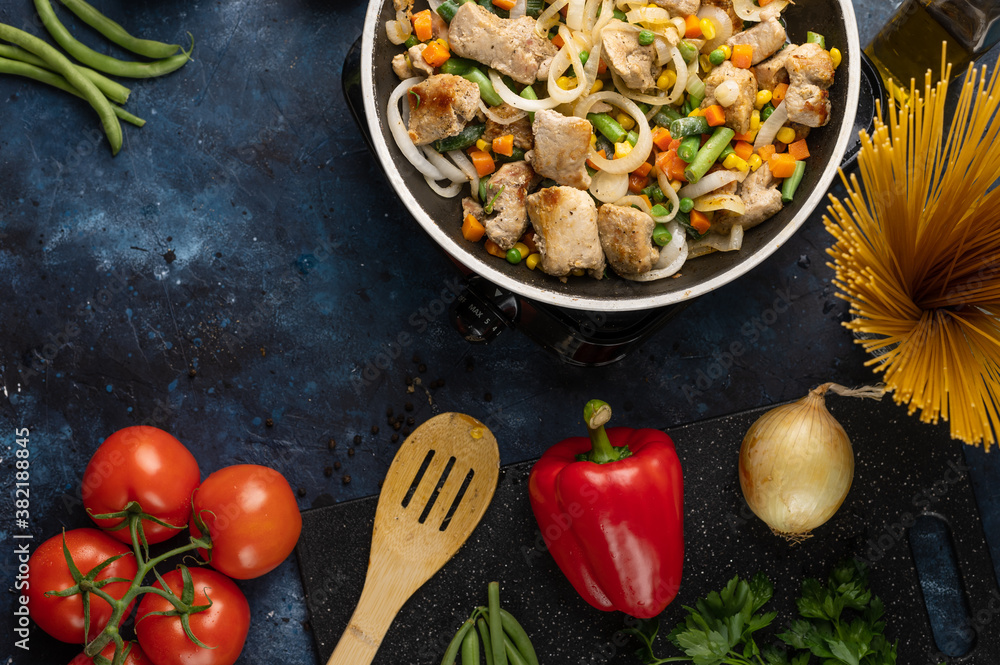 Top view of pan with frying chicken meat with vegetables for pasta on dark blue background. Professional kitchen table with wooden spatula, tomatoes, red pepper, onion and other ingredients for dish. Top view of pan with frying chicken meat with vegetables for pasta on dark blue background. Professional kitchen table with wooden spatula, tomatoes, red pepper, onion and other ingredients for dish.