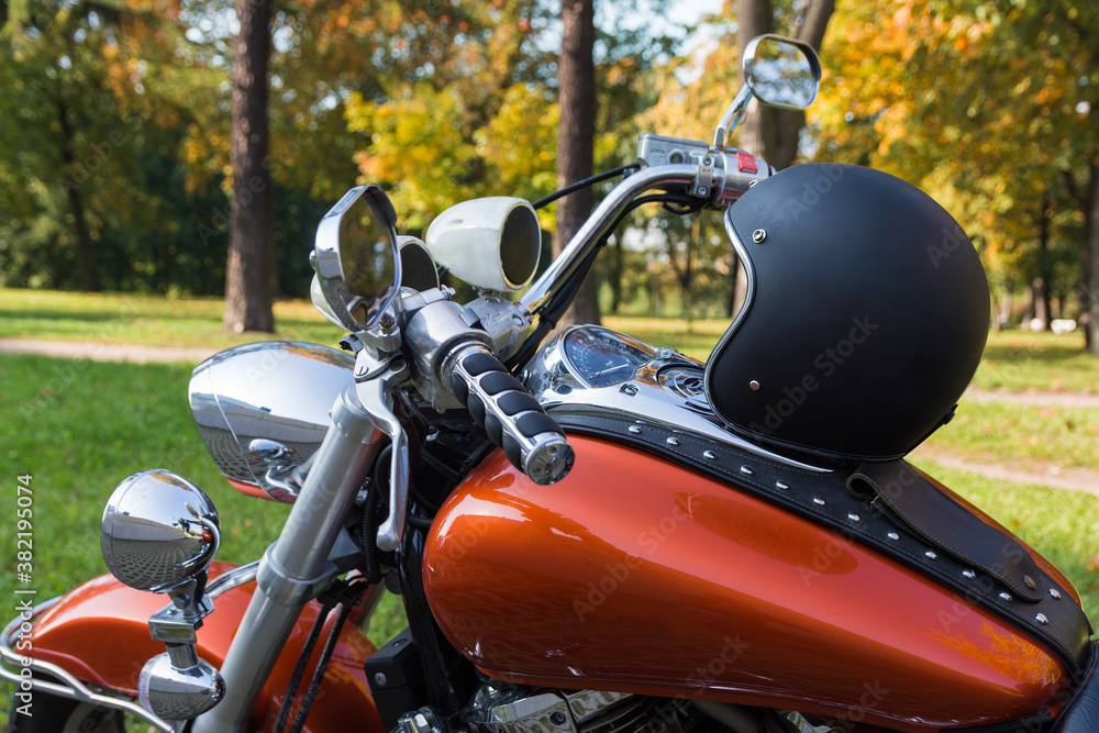 Photo of an open 3/4 black retro helmet, stand on the tank of the classic orange motorcycle. Motorcycle has lots of chrome details, music speakers and riveted tie, made of genuine brown leather. 