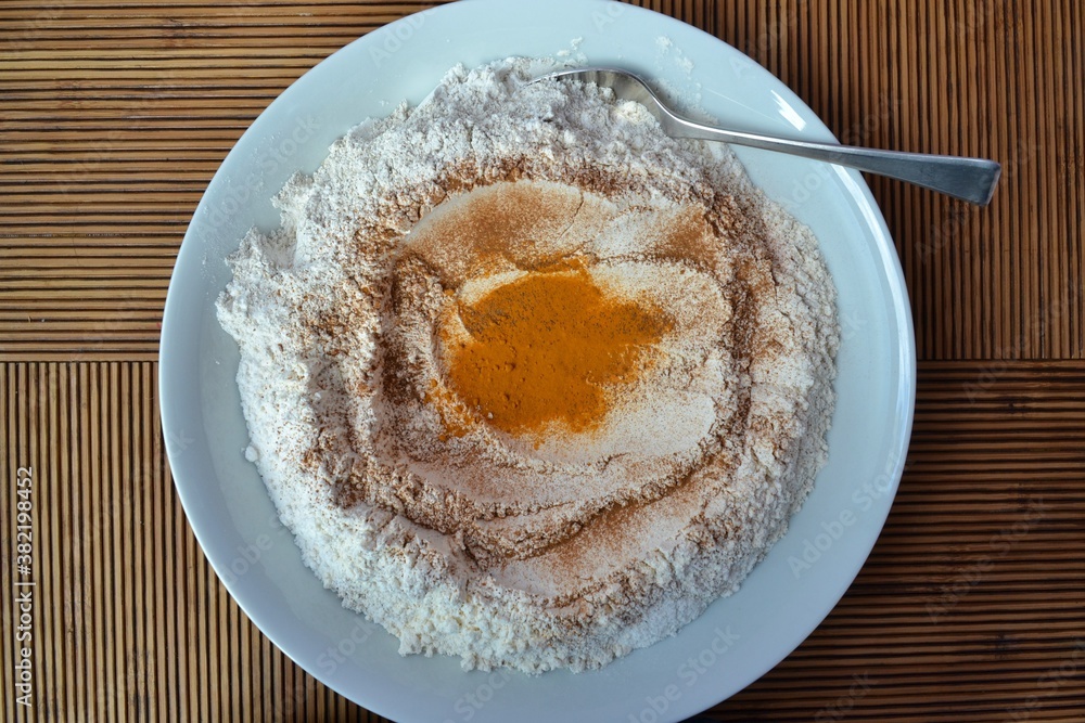 Plate of flour with spices like turmeric, paprika, cinnamon ... with a spoon to the side on a wooden striped table Plate of flour with spices like turmeric, paprika, cinnamon ... with a spoon to the side on a wooden striped table