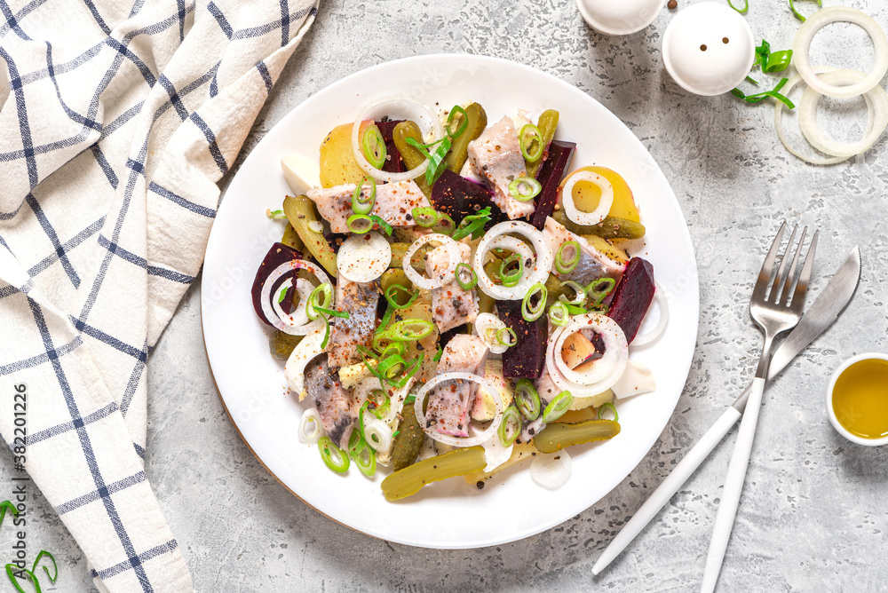 Salad of herring, beets, potatoes, eggs, pickling cucumbers and spices in white plate on gray concrete background top view.