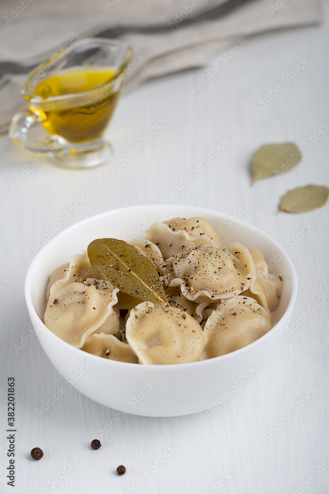 Homemade pelmeni or dumplings of Russian cuisine made of minced meat filling wrapped in dough served in white bowl with black pepper seasoning and bay leaf on white wooden background. Vertical