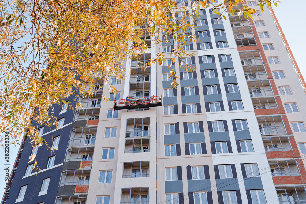 Fototapeta premium Finishing work concept. Workers are in a cradle and paint the facade of a new house under construction. Construction of new residential and office buildings.