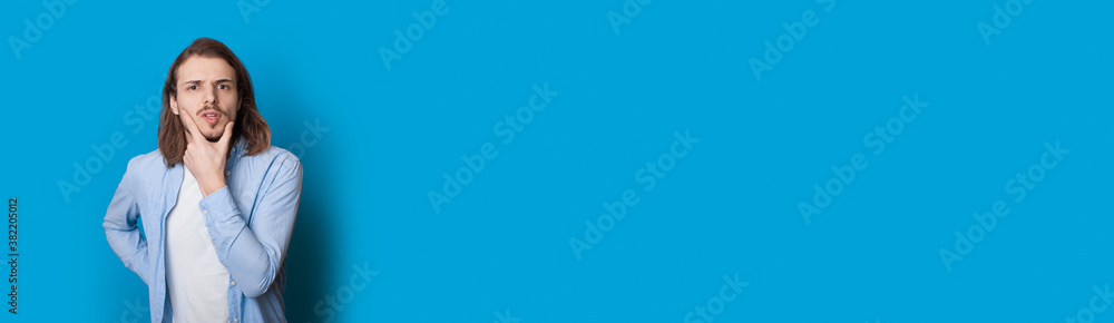 Confused caucasian man with long hair and bristle touching his chin with hand posing in casual clothes on a blue free spaced wall Confused caucasian man with long hair and bristle touching his chin with hand posing in casual clothes on a blue free spaced wall