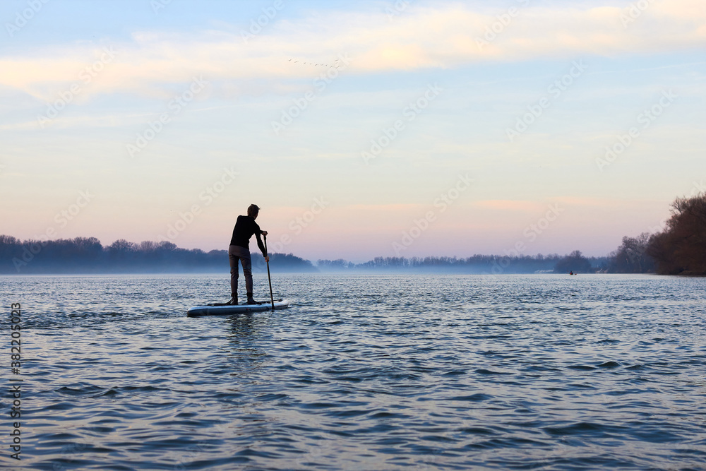 Teenager boy rowing on SUP (stand up paddle board) in autumn Danube river at foggy morning against the backdrop of autumn trees without leaves