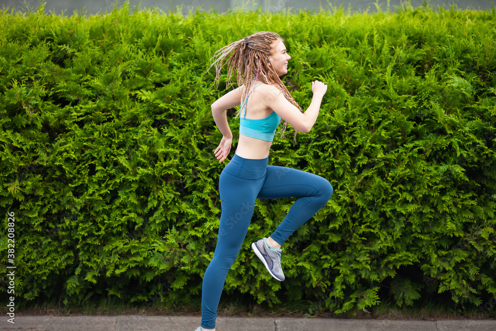 Young woman with dreadlocks running outdoors by the green fence. Sport and healthy lifestyle concept. .