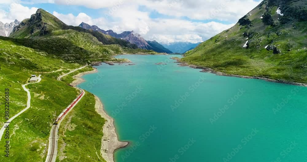 Red train passing near a turquoise blue lake in Switzerland at the bottom of a mountain. Aerial view with a drone of the beautiful landscape,under the blue sky 4K Red train passing near a turquoise blue lake in Switzerland at the bottom of a mountain. Aerial view with a drone of the beautiful landscape,under the blue sky 4K