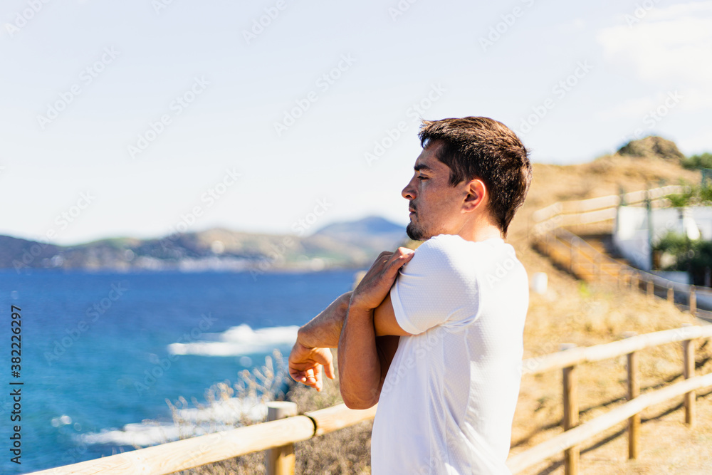 Young sportsman stretching arms outdoors while looking at the horizon with the sea to the side.