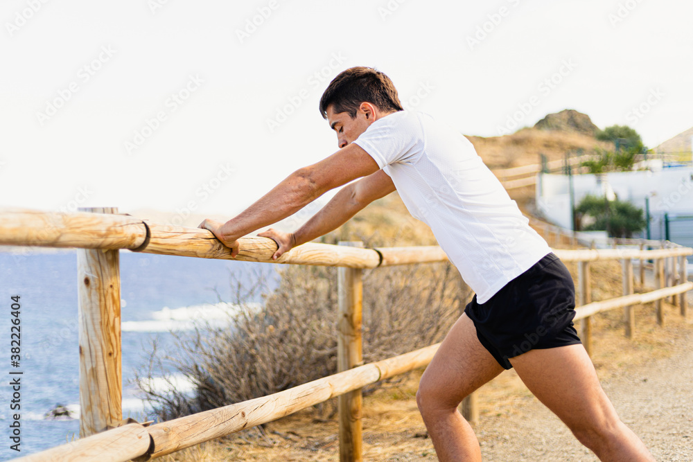 Young sportsman stretching his legs outdoors while looking at the horizon with the sea to the side.