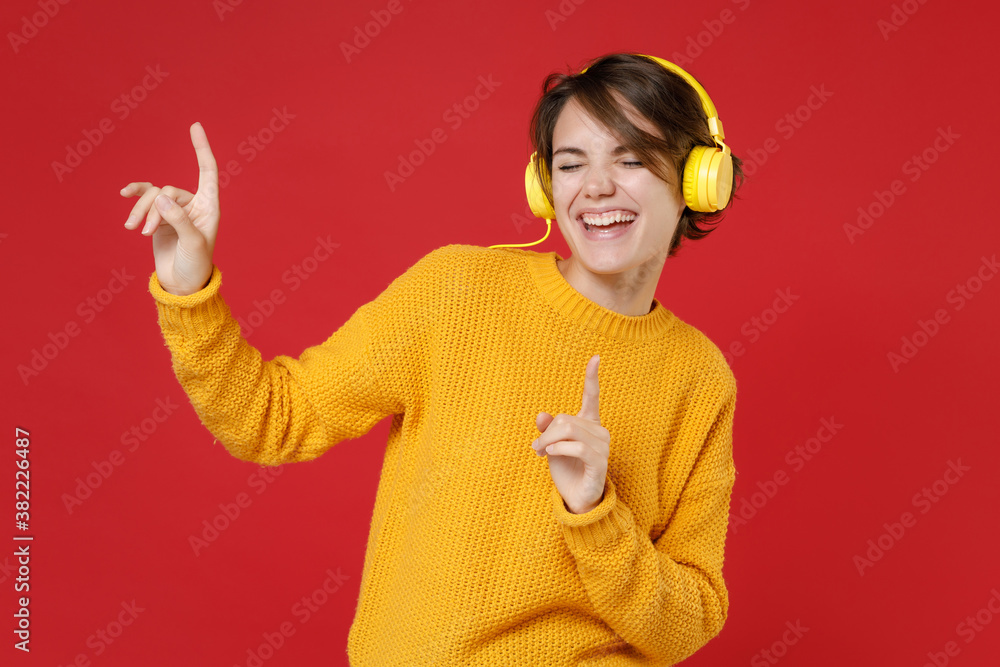 Funny pretty young brunette woman 20s in casual yellow sweater standing listening music with headphones dancing pointing index fingers up isolated on bright red colour background studio portrait. Funny pretty young brunette woman 20s in casual yellow sweater standing listening music with headphones dancing pointing index fingers up isolated on bright red colour background studio portrait.