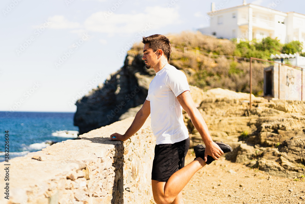 Young sportsman stretching his legs outdoors while looking at the horizon with the sea to the side.