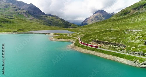 Aerial view with a drone of a red train passing near a blue lakeat the bottom of a mountain in Switzerland . Beautiful landscape under the blue sky 4K photo