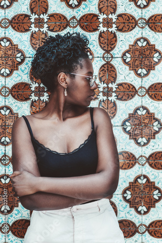 A vertical shot of a lovely young black female in a tank top and eyeglasses, with a curly Afro hair, standing in front of a tiled patterned Azulejo wall traditional for Portugal and looking aside © skyNext