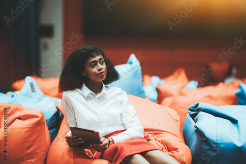 Portrait of a young beautiful African-American businesswoman sitting on an orange cushion with a tablet pc in hands indoors of a modern office chillout coworking area and thoughtfully looking aside photo