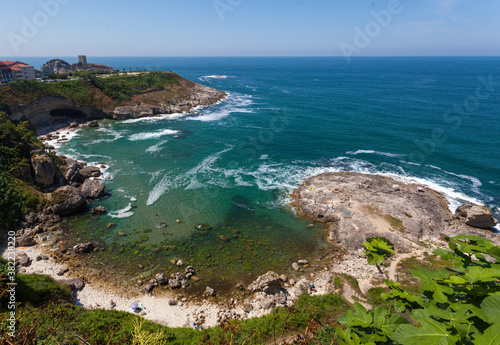 U shaped coast with lots of rocks inside. People are swimming in the sea while waves are hitting them. Castle is seen in a distance. people are lying over the rocks. In front, tree leaves are visible.