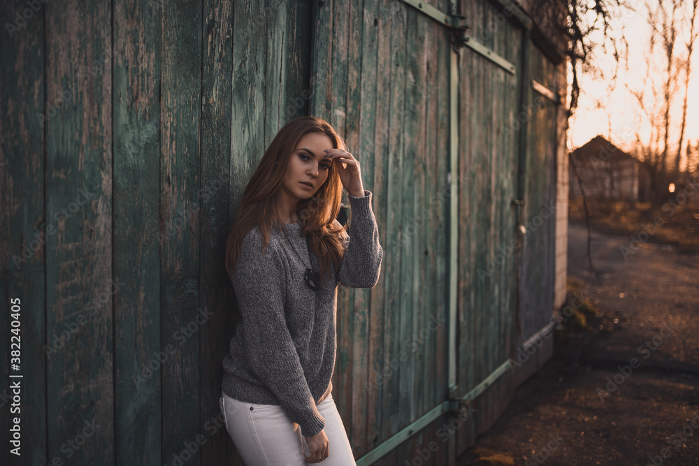 Fototapeta premium beautiful young blonde model girl. White pants. gray knitted sweater. black boots. wooden pendant on the neck in the form of a horse. On the Sunset. Portrait. 