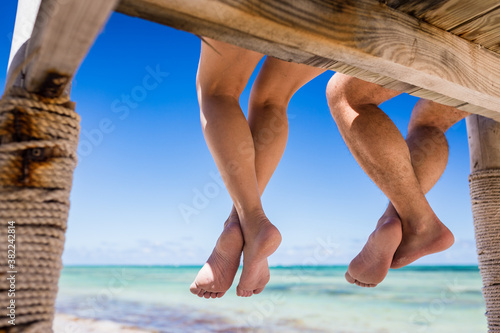 Fototapeta Naklejka Na Ścianę i Meble -  Close up view of legs of couple woman and man hanging from the wooden pier over the blue water of Caribbean Sea, Punta Cana, Dominican Republic 