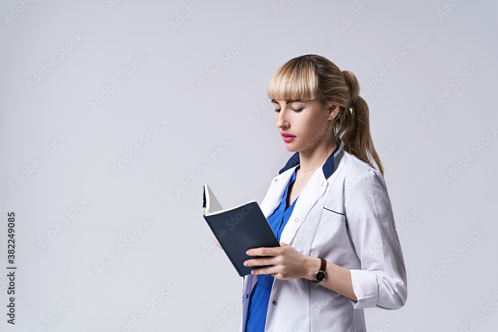 Confident young woman doctor standing with notebook or medical book in her hands. Cheks records, recipes or prescriptions. Intern in white medical clothes posing on light grey background. Confident young woman doctor standing with notebook or medical book in her hands. Cheks records, recipes or prescriptions. Intern in white medical clothes posing on light grey background.