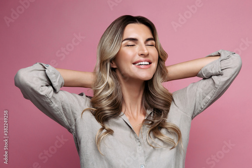 Studio shot of happy emotional woman in linen shirt smiling widely, showing perfect white teeth, closing eyes with pleasure, holding her hands behind head, dressed , isolated on pink background. 