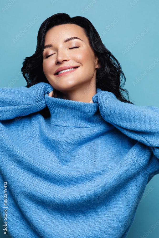 Delighted positive brunette woman smiling, wear high neck knitted blue sweater, has good mood, dreamily closed her eyes, isolated on studio blue background. Winter season. Delighted positive brunette woman smiling, wear high neck knitted blue sweater, has good mood, dreamily closed her eyes, isolated on studio blue background. Winter season.