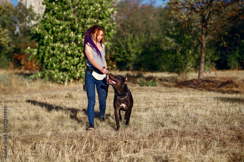 young informal woman with piercings and pigtails walks in the park with her dog, black pit bull on a walk