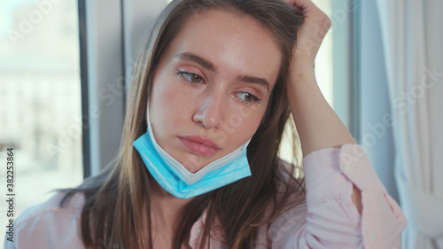 Sad caucasian woman wearing a surgical mask sitting by window at home. Beautiful girl is upset because of coronavirus outbreak. Self-isolation. Lockdown stress.