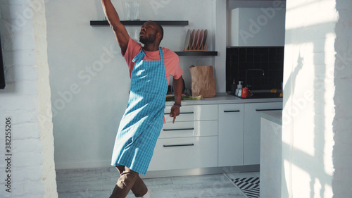 Charismatic and joyful afro-american man chef wearing cooking apron dancing in the kitchen. Funny excited guy having fun alone at home. Entertainment. Casual people.