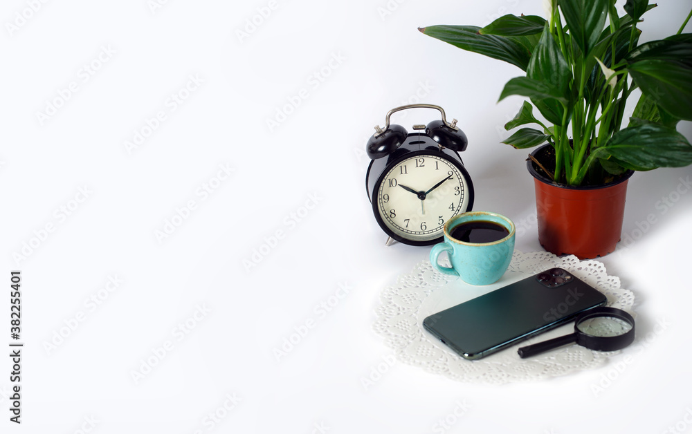 Cup of black coffee with alarm clocks, magnifier, phone and Spathiphyllum green houseplant on a white background. Side view with copy space for your text. Studio shot. Coffee time in the morning Cup of black coffee with alarm clocks, magnifier, phone and Spathiphyllum green houseplant on a white background. Side view with copy space for your text. Studio shot. Coffee time in the morning