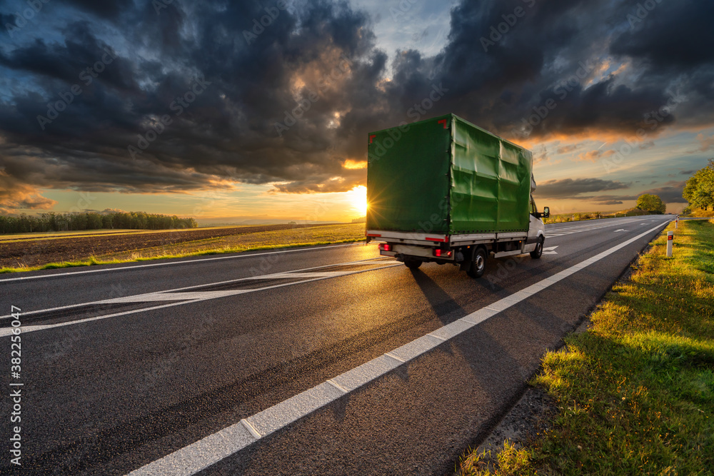 Delivery van driving on the asphalt road in rural landscape in the rays of the sunset with dark cloud