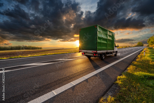 Obraz na płótnie Delivery van driving on the asphalt road in rural landscape in the rays of the sunset with dark cloud w salonie