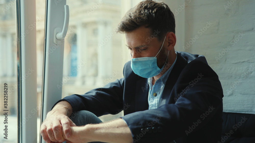 Sad businessman remove medical mask at home sitting on windowsill and looking at street on quarantine COVID-19 pandemic epidemic depressed portrait slow motion