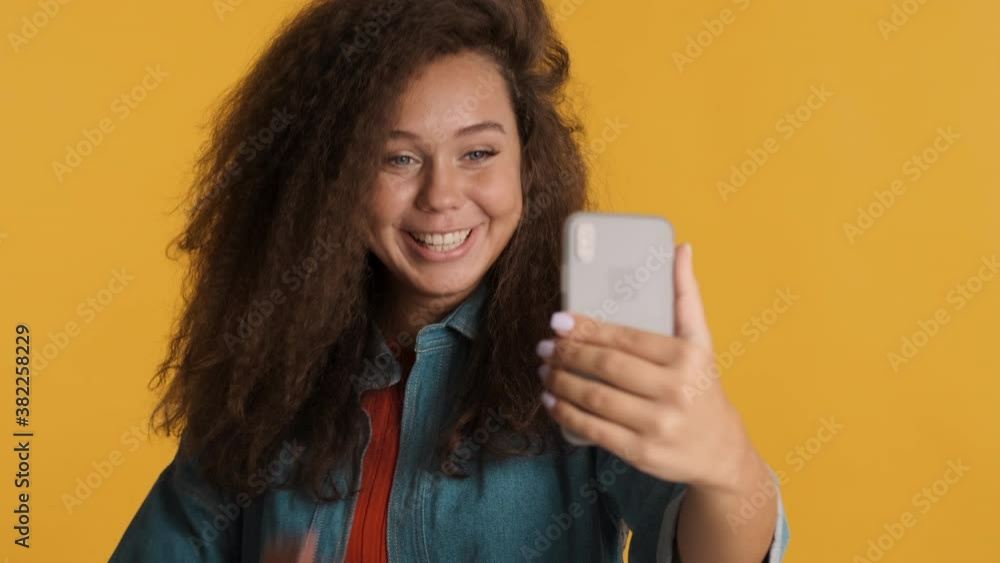 Beautiful brunette girl making video call happily sending air kiss to boyfriend talking with him on smartphone isolated on yellow background Beautiful brunette girl making video call happily sending air kiss to boyfriend talking with him on smartphone isolated on yellow background