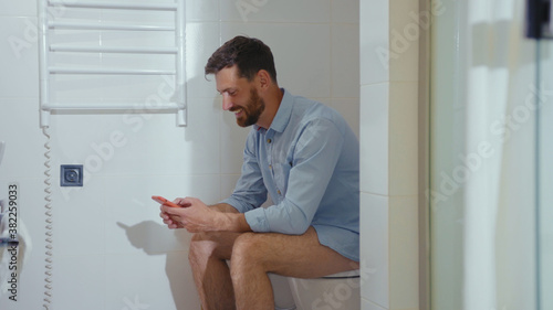 Happy businessman use phone sitting on the toilet smiling mobile restroom smartphone internet home bathroom close up slow motion