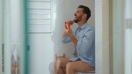 Happy businessman use phone sitting on the toilet smiling mobile restroom smartphone internet home bathroom close up slow motion