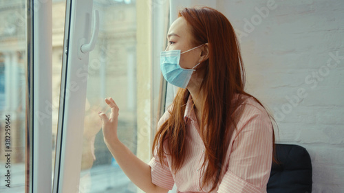 Upsed asian woman in medical mask at home sitting on windowsill and looking at street on quarantine COVID-19 pandemic epidemic depressed portrait slow motion