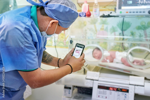 A father in protective clothes taking a cell phone of his newborn baby in an incubator