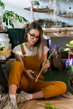Small business. Florist woman surrounded by tropical plants cutting the stem of irises flowers using secateurs, sitting on the floor in flower store.