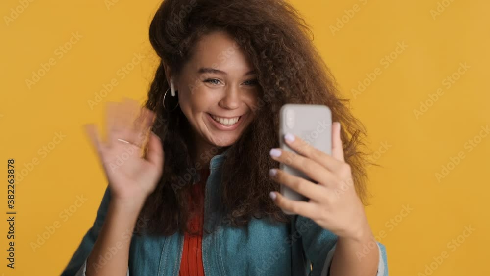 Charming brunette girl happily making video call to friend waving palm in hello gesture using smartphone isolated on yellow background Charming brunette girl happily making video call to friend waving palm in hello gesture using smartphone isolated on yellow background