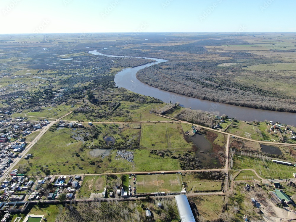 Foto aérea de los suburbios de una ciudad, con vista parcial de un río serpenteante, con vegetación en la costa. 