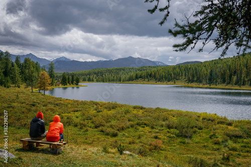 Couple in hooded jackets sitting on a bench, backs to the camera against the backdrop of beautiful lake in the mountains. Guy and girl resting, admiring the landscape. Travel, outdoor activities.