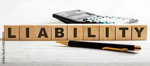 The word LIABILITY is written on the wooden cutouts between the calculator and the pen on a light background.