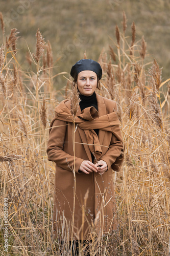 Portrait of a beautiful young model in a black shiny beret and a beige wool coat smilling, posing on field in sunny autumn day . 