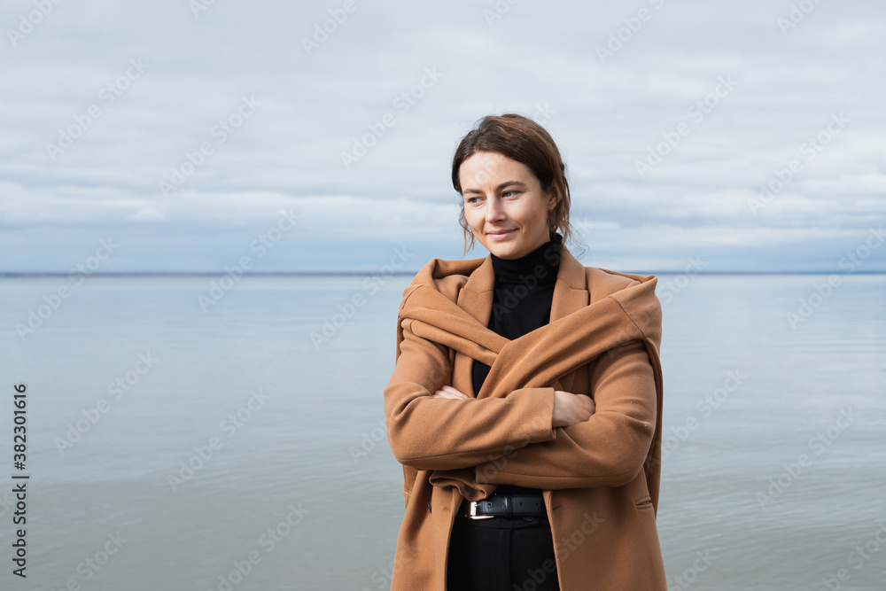 Fashion lifestyle portrait of young trendy woman dressed in  beige wool coat smiling, walking on the beach,  on background a sea .Portrait of joyful woman