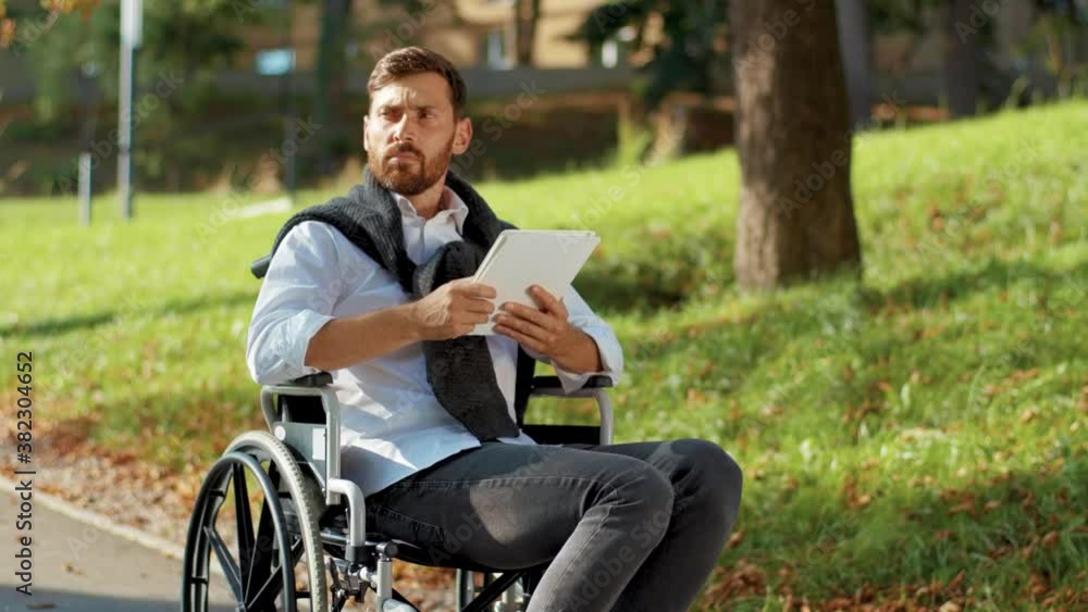 Serious young man on a wheelchair with tablet computer in park at sunlight disability medicine. Possibility handicapped handicap tablet internet. Slow motion Serious young man on a wheelchair with tablet computer in park at sunlight disability medicine. Possibility handicapped handicap tablet internet. Slow motion