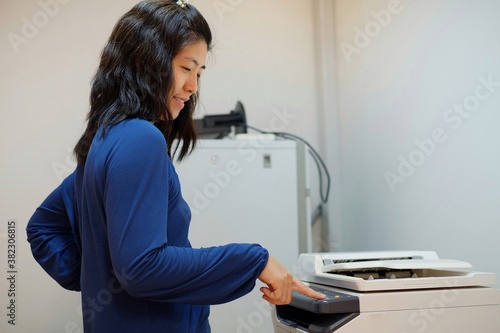 A middle aged Asian woman, working as a secretary is copying documents using a multifunction laser printer at her office.