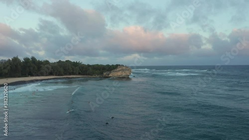 Surfers at sunset in Hawaii drone shot over the waves overlooking bay and beach. Slow motion aerial view flying backwards revealing surfing spot. Poipu Kauai photo