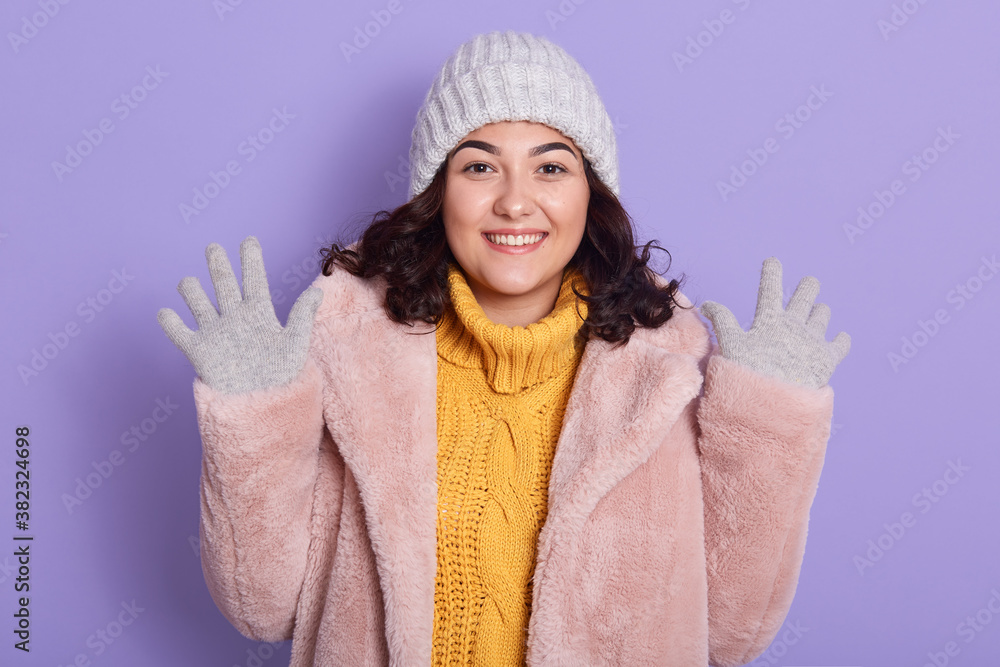Happy brunette Caucasian woman spreads hands up in air, has good mood, wears warm coat, sweater cap and mittens, looks at camera with toothy smile, poses indoor against lilac wall. Happy brunette Caucasian woman spreads hands up in air, has good mood, wears warm coat, sweater cap and mittens, looks at camera with toothy smile, poses indoor against lilac wall.
