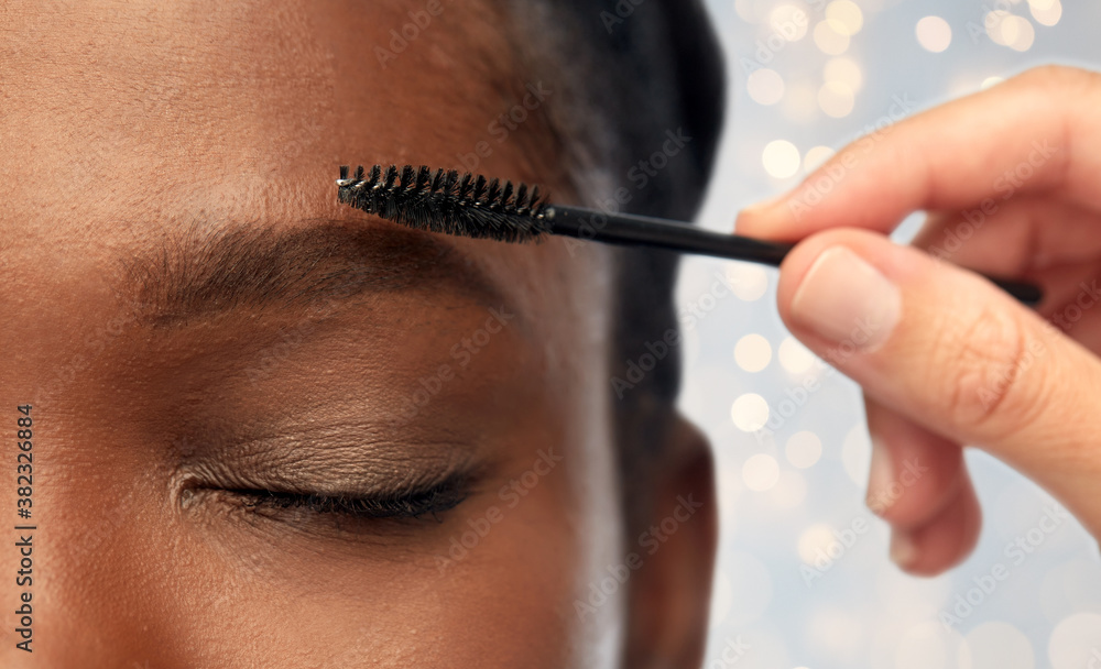beauty, make up and cosmetics concept - close up of face of young african american woman and hand with mascara brush applying eyebrow shadows over festive lights background