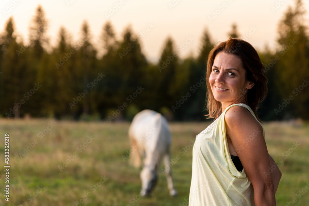 Obraz premium Beautiful woman at horse farm with white horse in the background at sunset. Golden hour photography