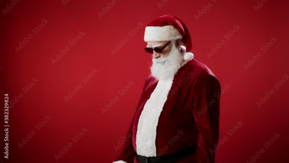 Lockdown of Santa Claus putting on fashionable red sunglasses standing against red background then looking at camera and making cool gesture with his fingers Lockdown of Santa Claus putting on fashionable red sunglasses standing against red background then looking at camera and making cool gesture with his fingers