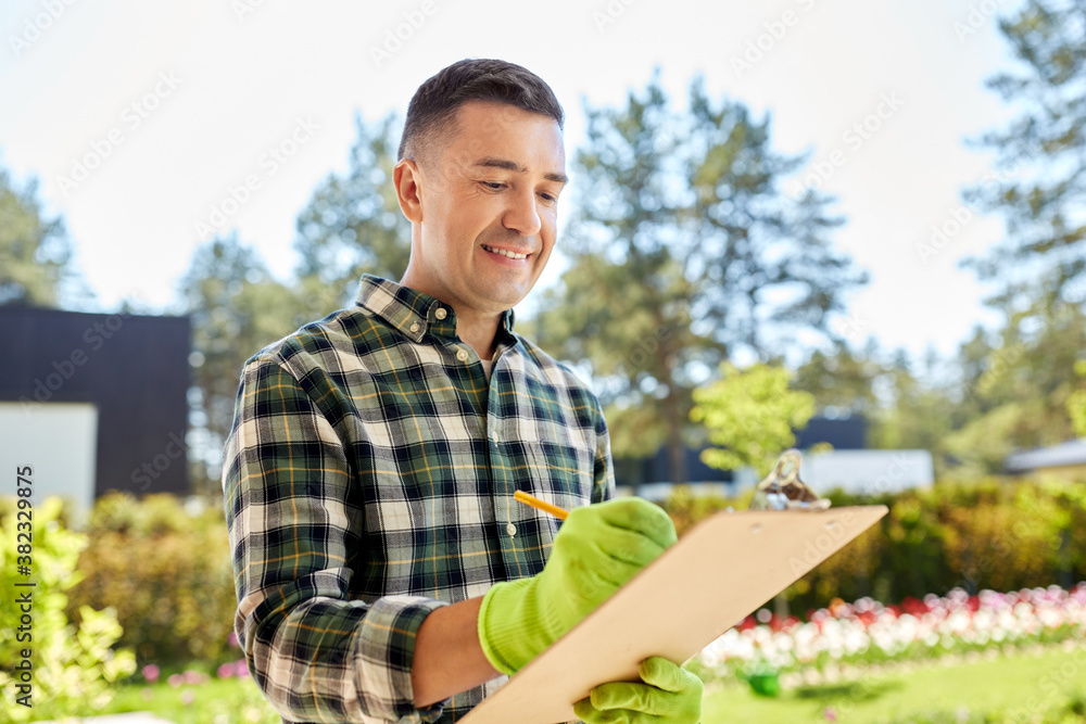 Fototapeta premium gardening and people concept - happy smiling middle-aged man in gloves writing to clipboard at summer garden